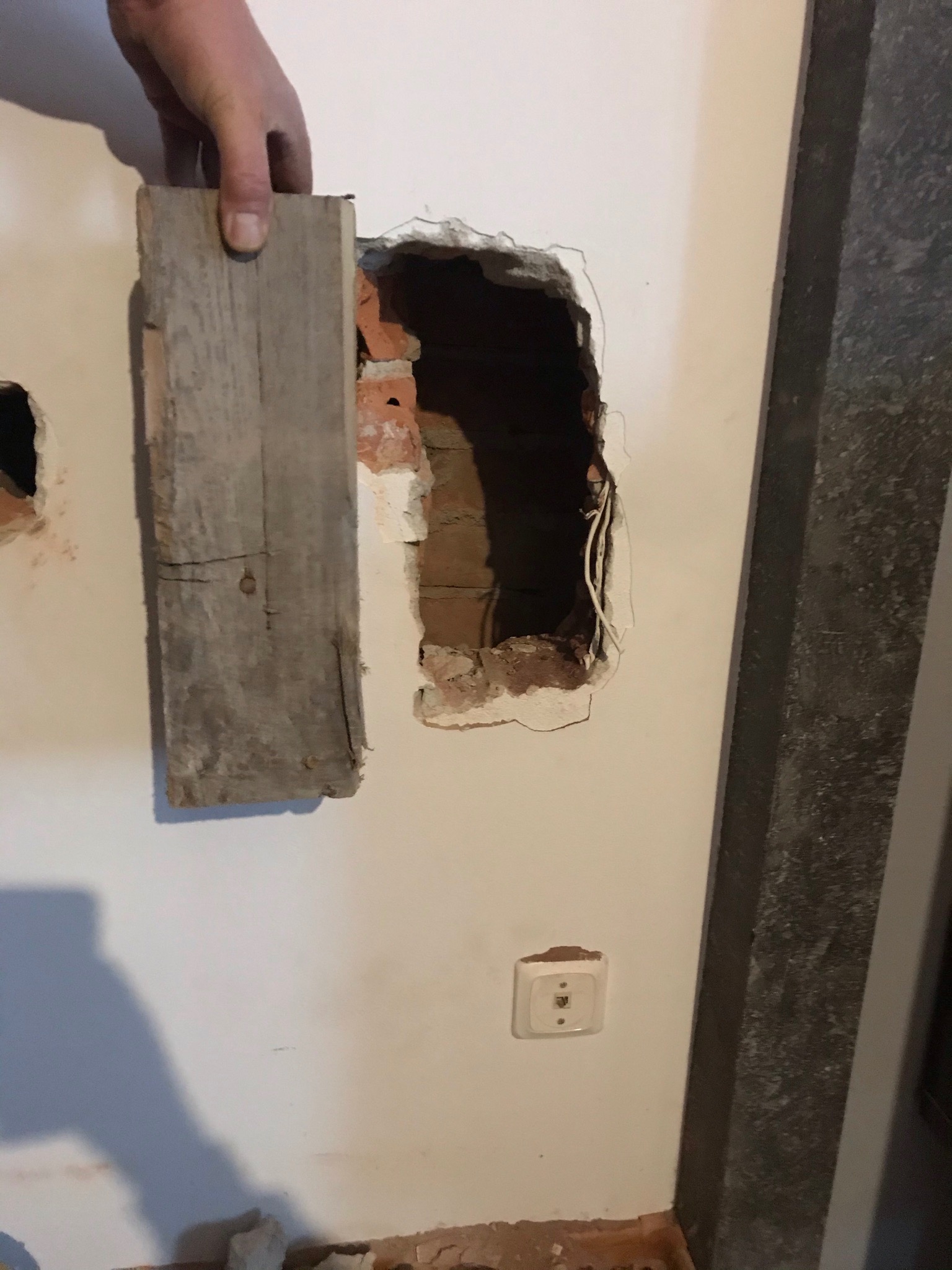 Fragment ściany z odsłoniętą cegłą i instalacją elektryczną, zakrywany drewnianą deską, widoczny gniazdko telefoniczne.