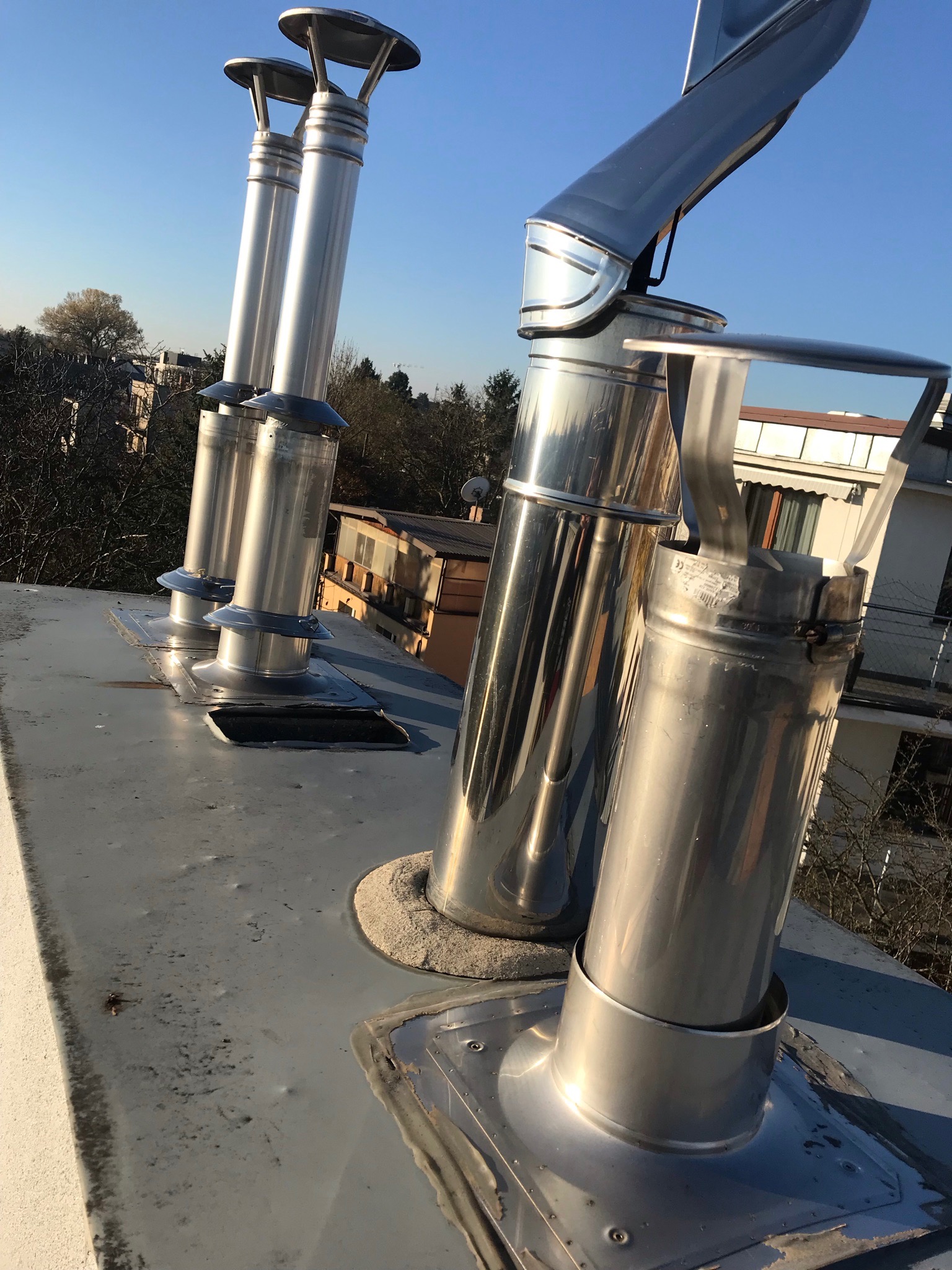 Błyszczące, metalowe kominy wentylacyjne na szarym dachu, widoczne z bliska na tle błękitnego nieba i zabudowań.