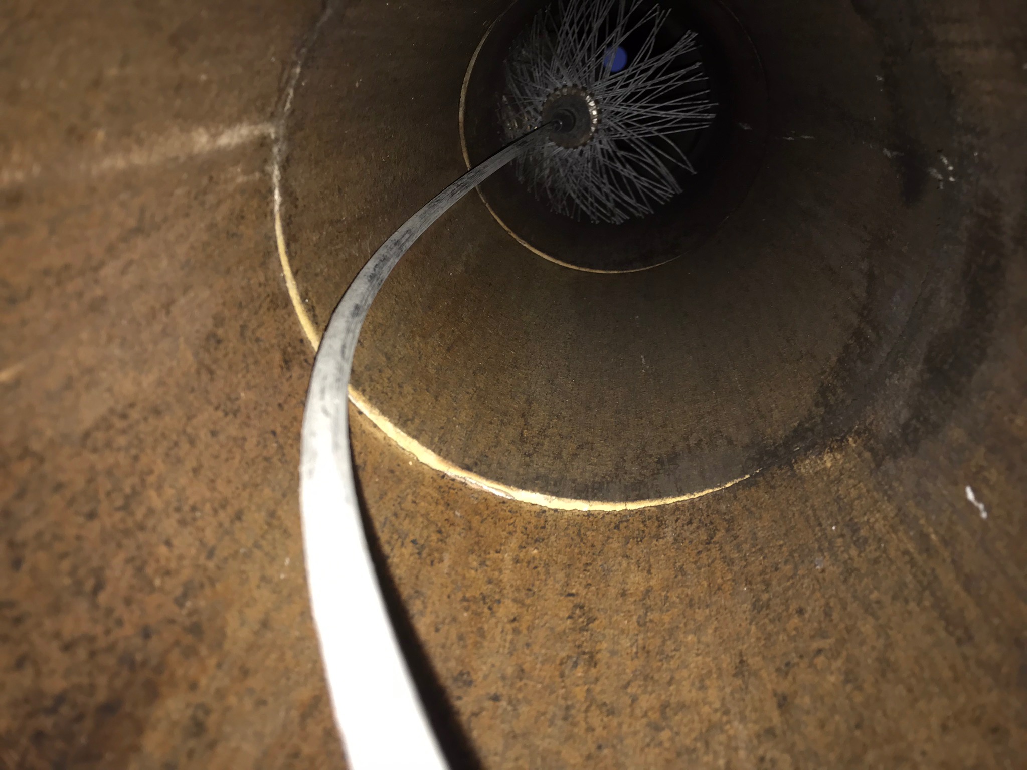 Wnętrze zardzewiałego komina, widok z dołu na szczotkę kominiarską z szarymi włoskami i niebieskim elementem mocującym, podłączoną do elastycznego przewodu.