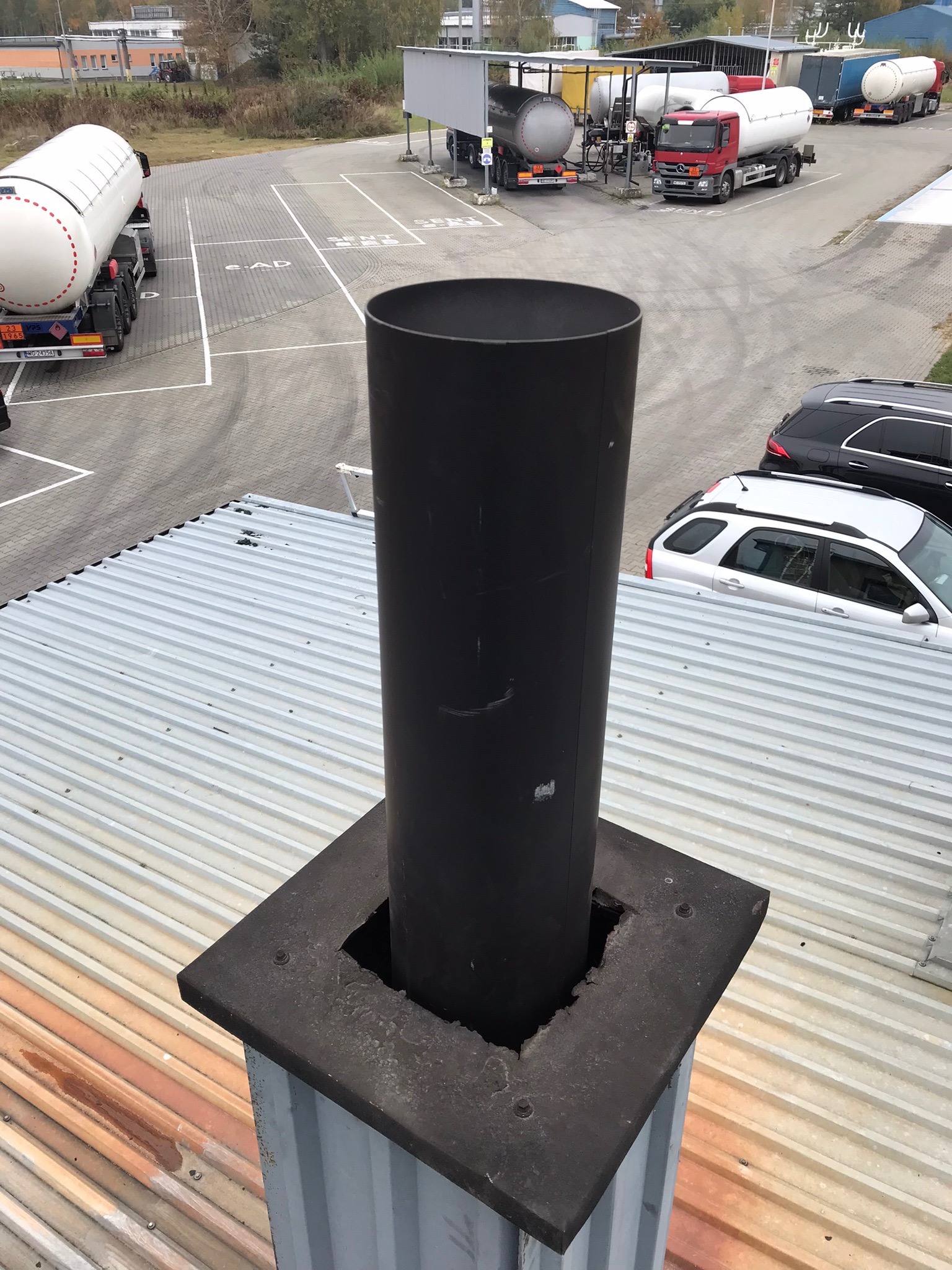 Czarny komin z rurą na dachu z blachy falistej, widok z góry, w tle parking z cysternami i samochodami.