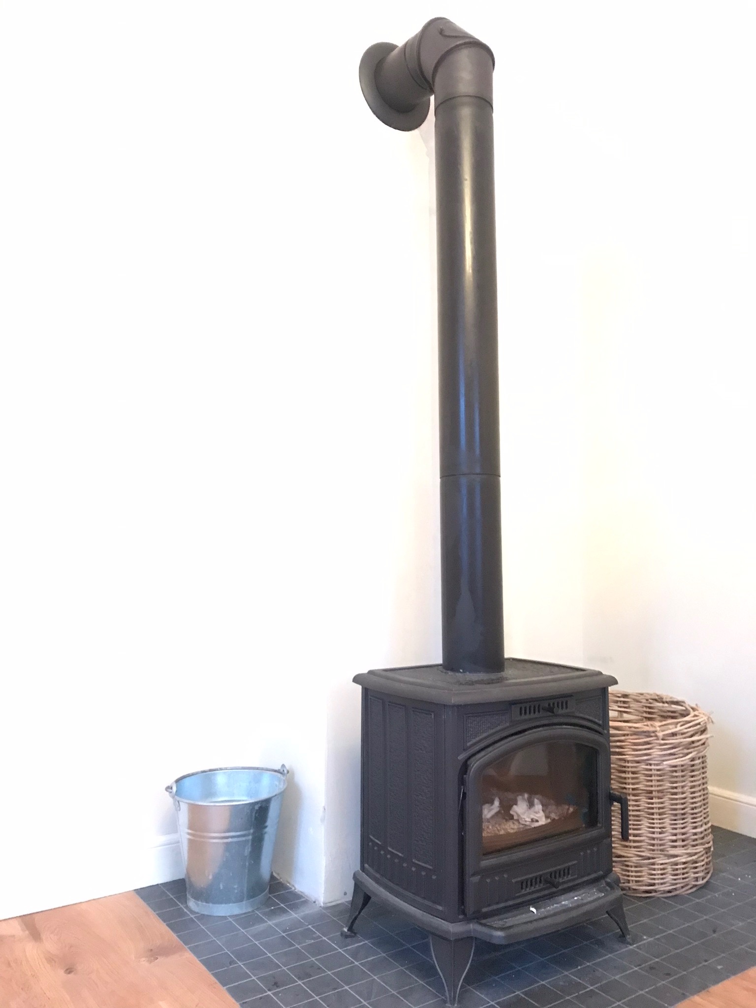 Czarny piec wolnostojący z rurą odprowadzającą dym, obok ocynkowane wiadro i wiklinowy kosz na tle białej ściany i podłogi w szarą szachownicę.
