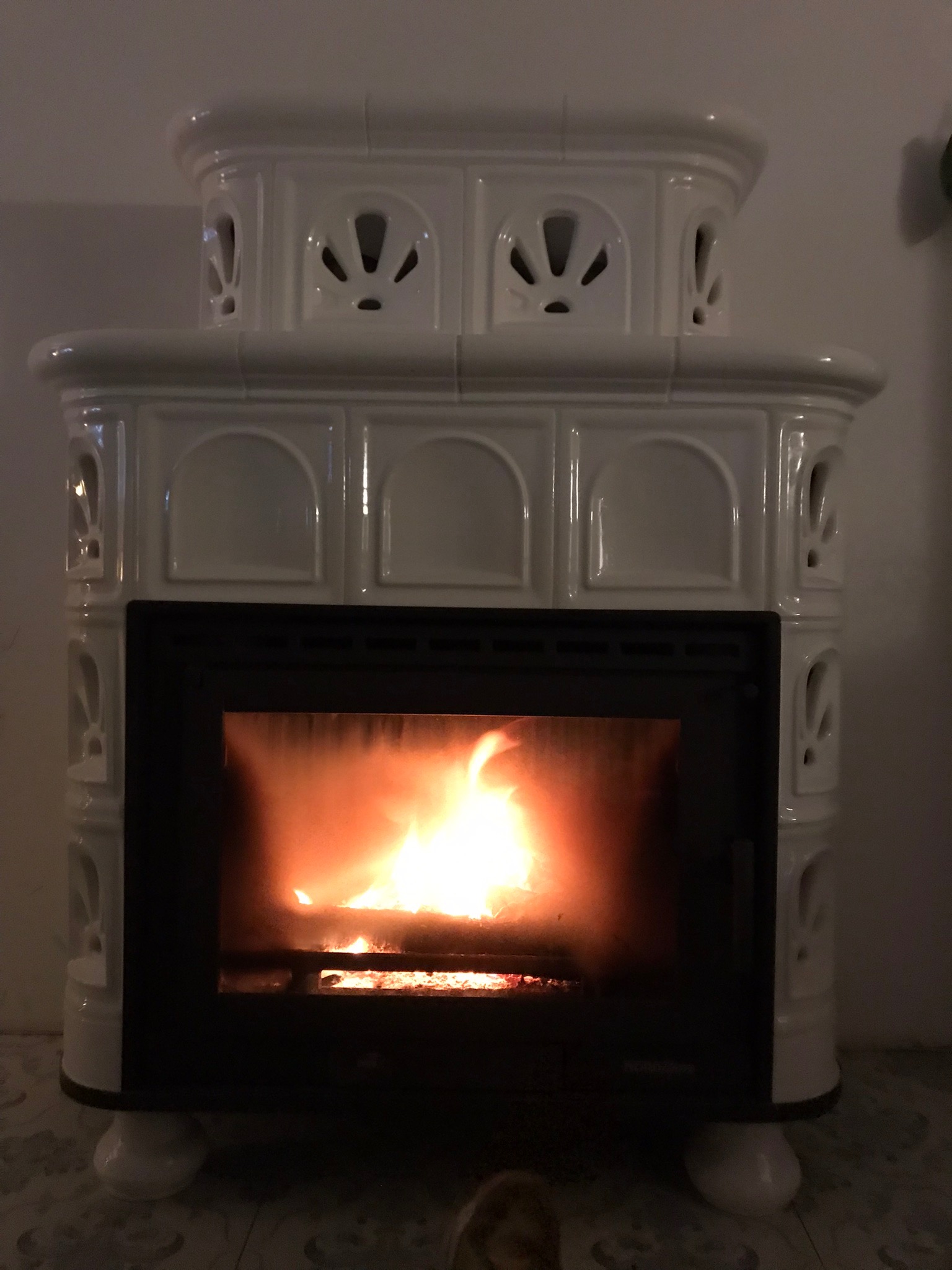 Biały, kaflowy piec z palącym się wewnątrz ogniem, widoczne dekoracyjne otwory wentylacyjne w górnej części.