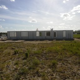 Robert Popiela - Surowy stan budynku w trakcie budowy, widoczne szare ściany z otworami okiennymi na tle nieba i pola z zielenią.