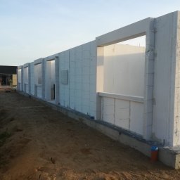 Robert Popiela - Wznoszenie ściany domu z białych bloczków betonowych, widoczne otwory okienne i drzwiowe, teren budowy w tle.