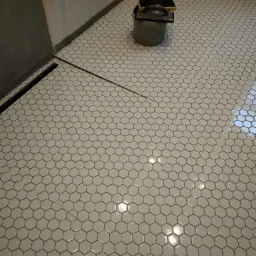 Ułożona podłoga z białych, heksagonalnych płytek w łazience, z widocznym odpływem liniowym, wiadro z narzędziami glazurniczymi i opakowaniem po kleju w tle.