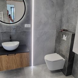 Robert Popiela - Nowoczesna łazienka z szarymi płytkami imitującymi marmur, okrągłe lustro z podświetleniem LED, podwieszana toaleta i umywalka nablatowa na szafce z lamelami.