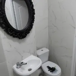 Nowoczesna łazienka z białymi marmurowymi płytkami, umywalką na postumencie i toaletą z deską z motywem kamieni, ozdobne czarne lustro w owalnej ramie.