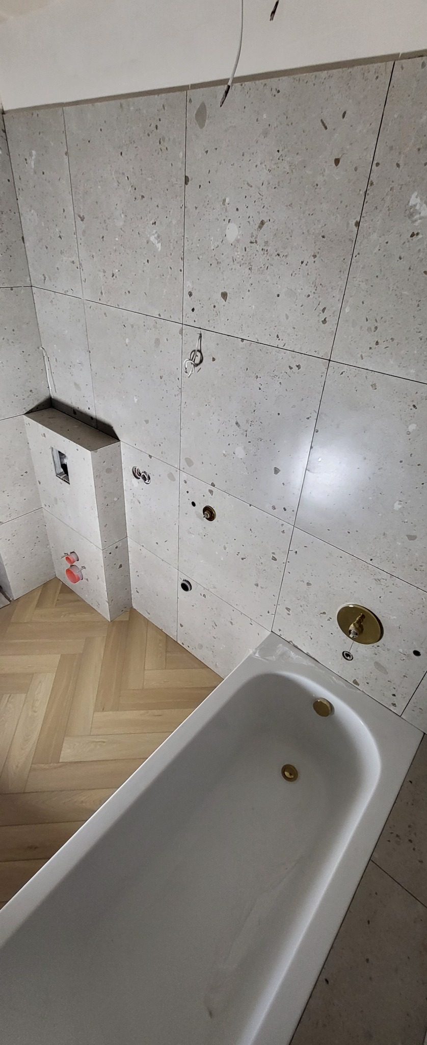 Biała wanna w łazience w trakcie remontu, widoczne płytki imitujące lastryko na ścianach, armatura w kolorze złotym oraz parkiet ułożony w jodełkę.
