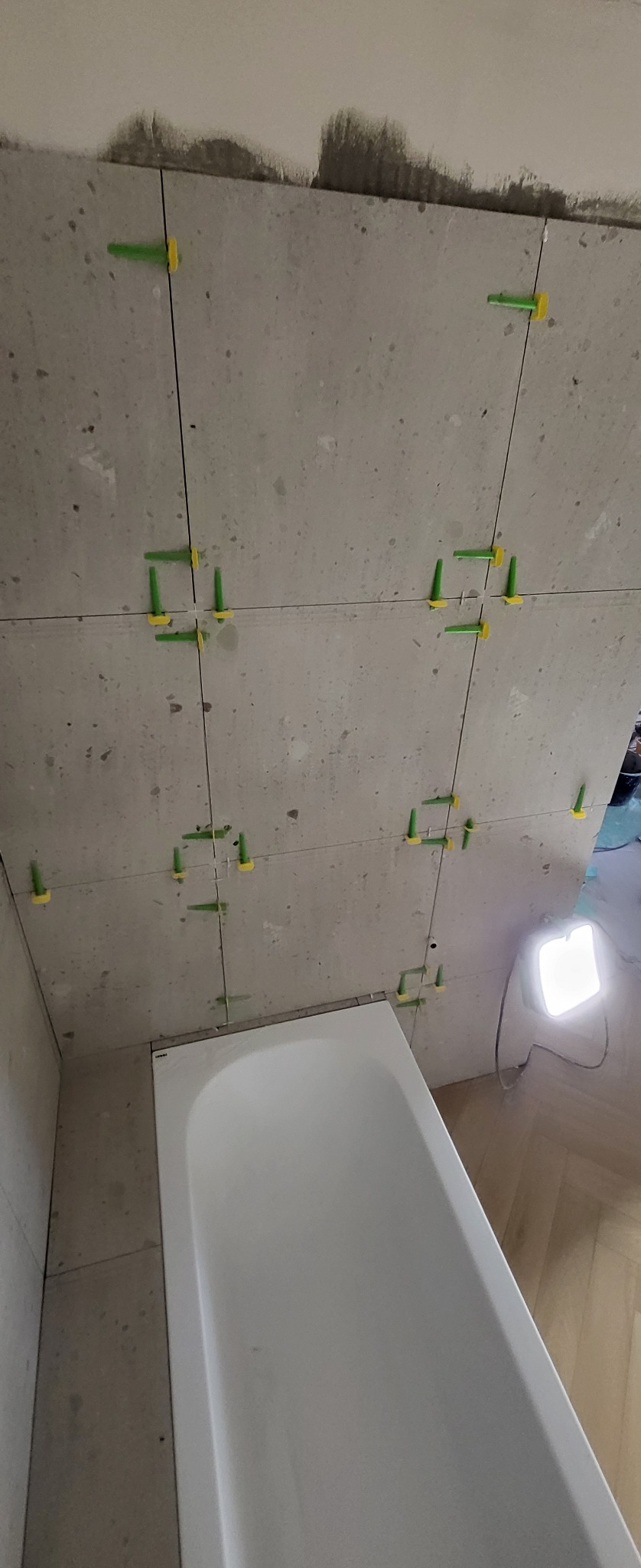 Świeżo ułożone, szare płytki w łazience z systemem poziomowania płytek (zielono-żółte kliny) i białą wanną w dolnej części kadru, oświetlone lampą budowlaną.