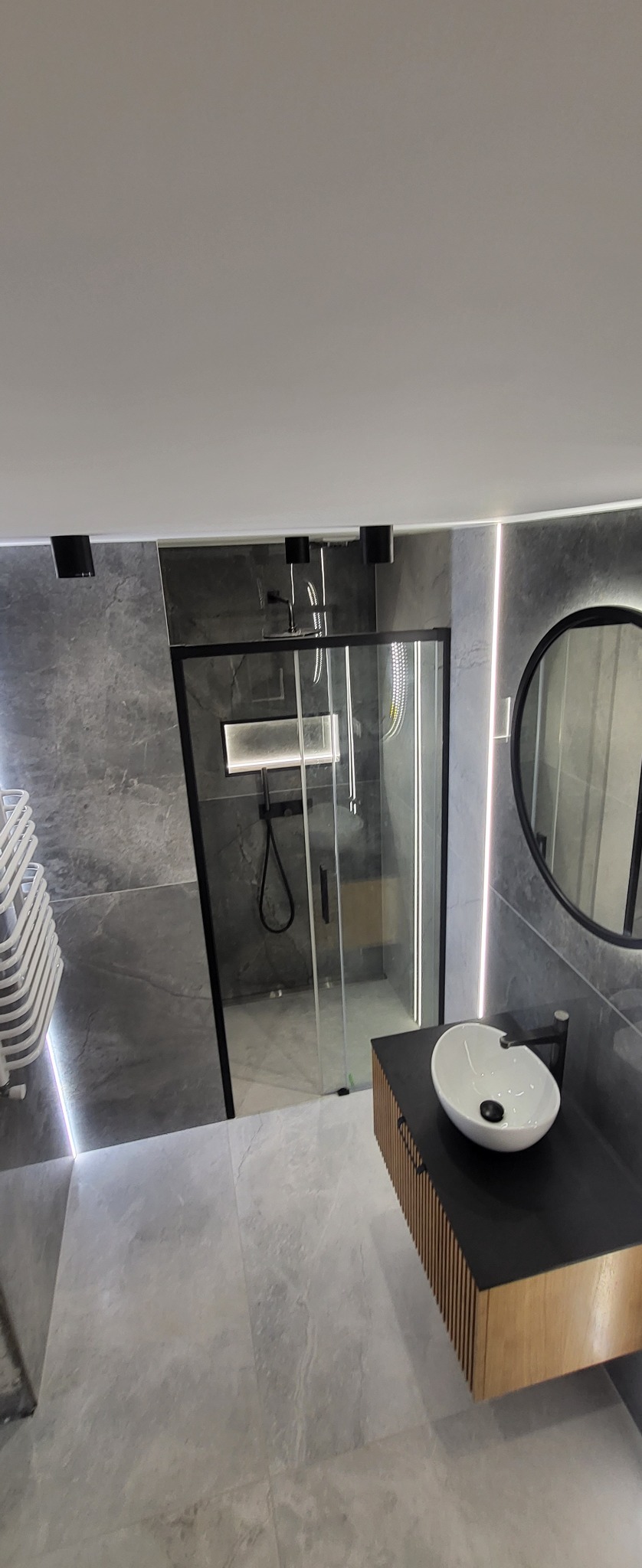Nowoczesna łazienka w szarościach z kabiną prysznicową typu walk-in, umywalką nablatową na szafce z drewnianymi lamelami i okrągłym lustrem z podświetleniem LED.