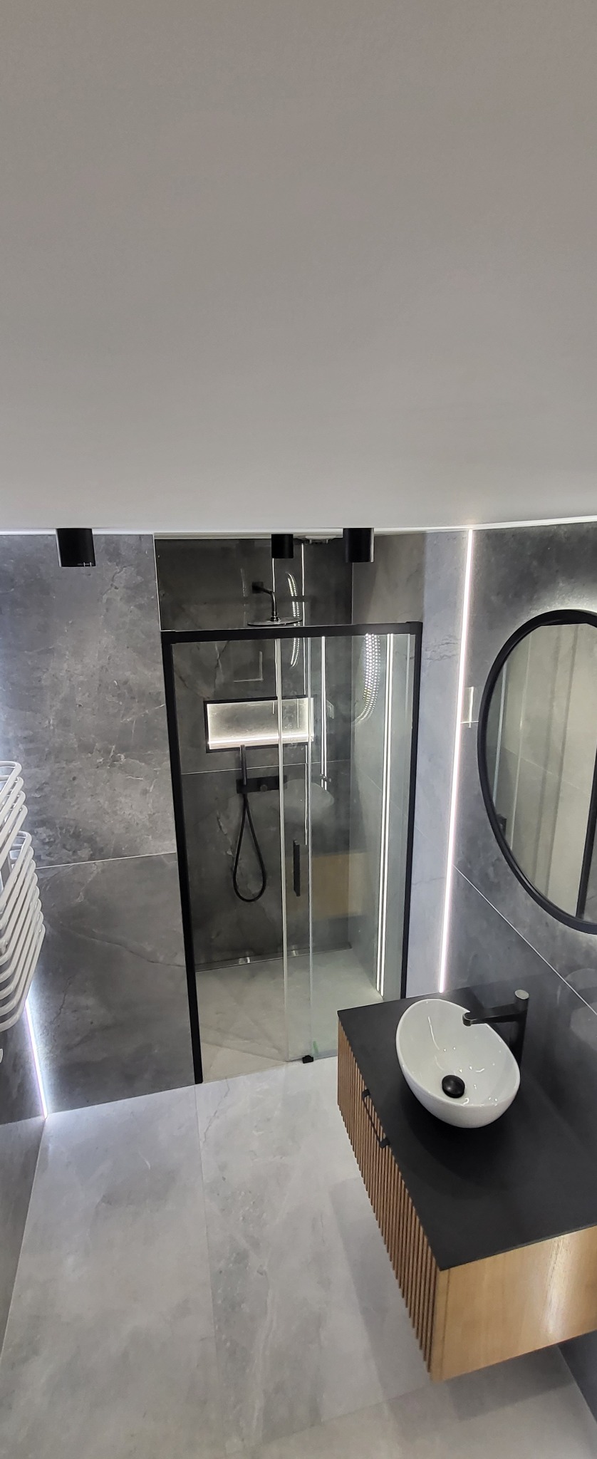 Nowoczesna łazienka z szarą marmurową płytką, kabiną prysznicową typu walk-in z czarnymi ramami, okrągłym lustrem z czarną obwódką i wiszącą umywalką z drewnianymi lamelami.