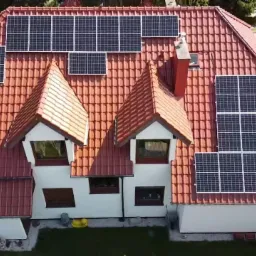 Widok z góry na dom z pomarańczową dachówką, na której zamontowano panele słoneczne w różnych konfiguracjach, w tym nad oknami dachowymi i szczytami dachu.