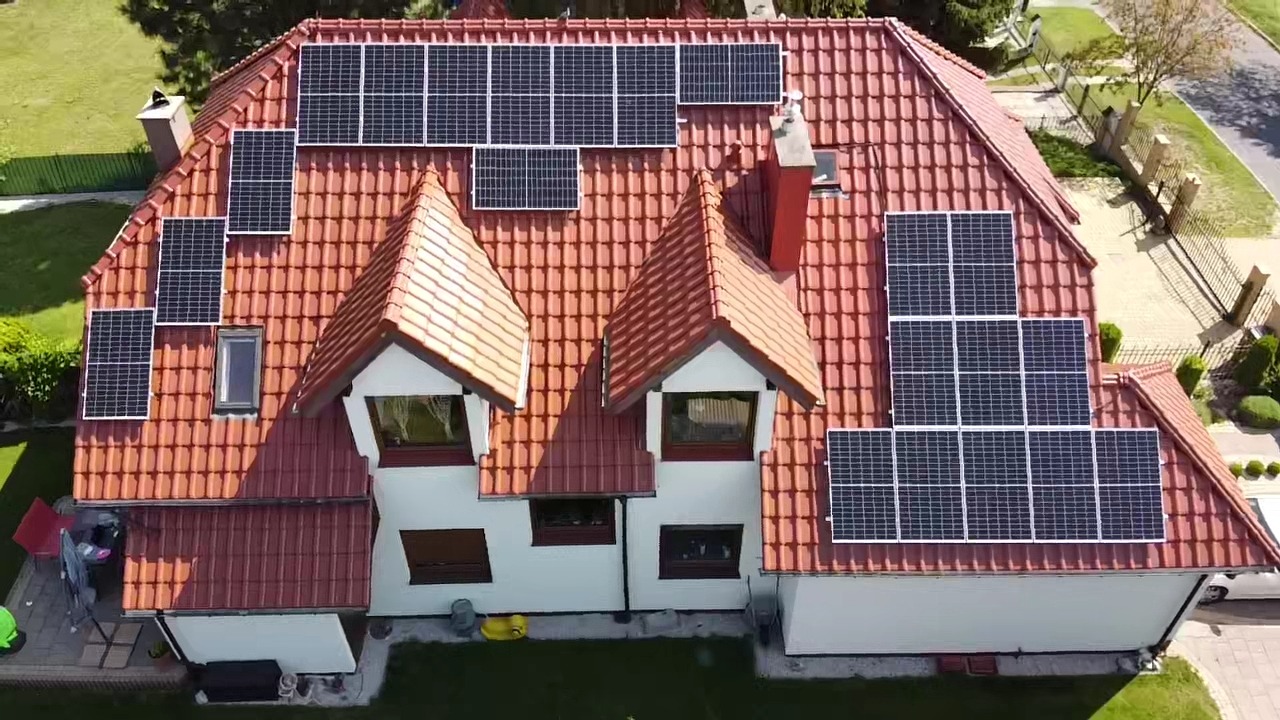 Widok z góry na dom z pomarańczową dachówką, na której zamontowano panele słoneczne w różnych konfiguracjach, w tym nad oknami dachowymi i szczytami dachu.