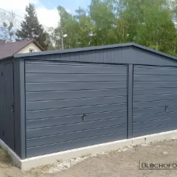 Szary, dwustanowiskowy garaż blaszany z drzwiami bocznymi, umieszczony na betonowej podmurówce, z widocznym logo producenta w prawym dolnym rogu.