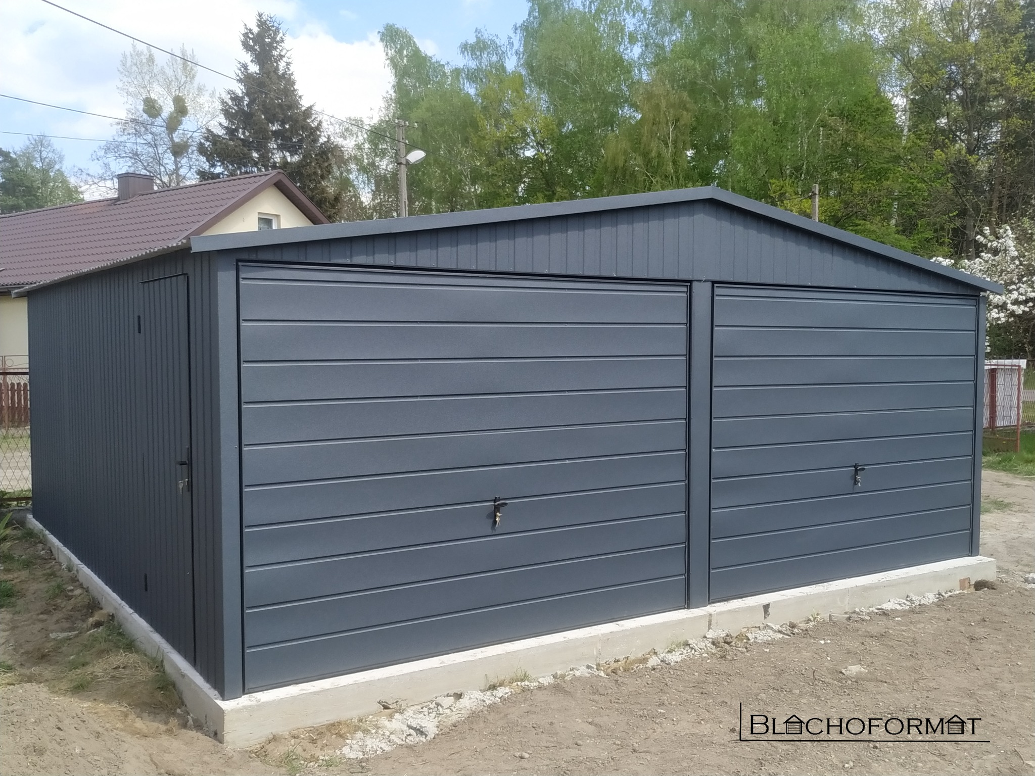 Szary, dwustanowiskowy garaż blaszany z drzwiami bocznymi, umieszczony na betonowej podmurówce, z widocznym logo producenta w prawym dolnym rogu.