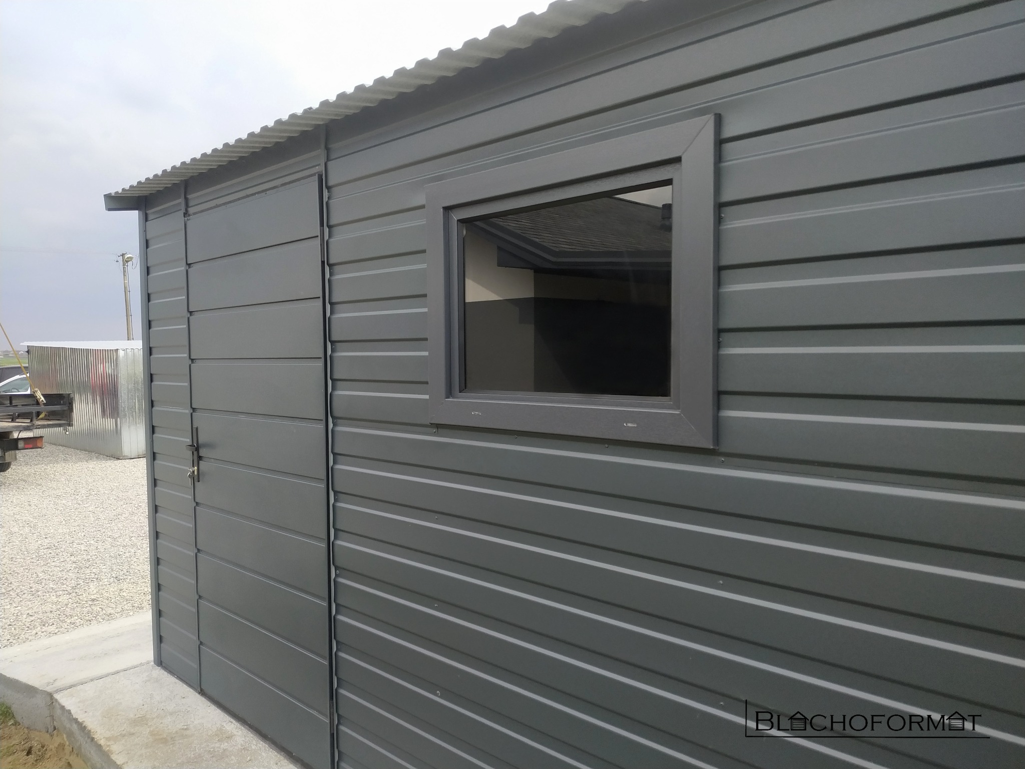 Szary garaż blaszany z oknem i drzwiami, widok z boku, zamek z kluczem w drzwiach, tło częściowo rozmyte.