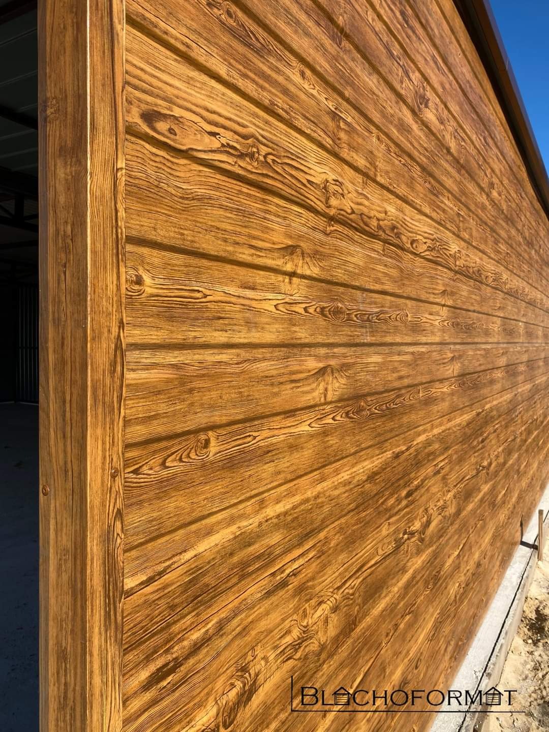 Zewnętrzna ściana garażu z blachy trapezowej imitującej drewno, widoczna z boku, z fragmentem niebieskiego nieba w tle.