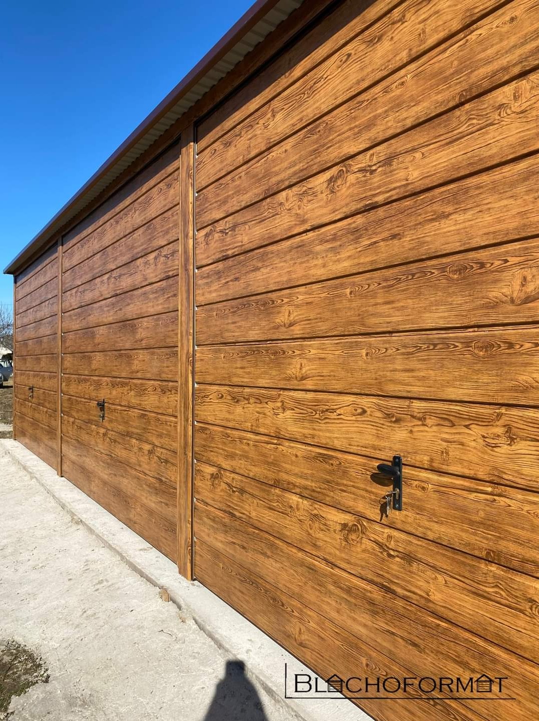 Długi, brązowy garaż blaszany z imitacją drewna, widoczny z boku, z kluczem w zamku, na tle błękitnego nieba.