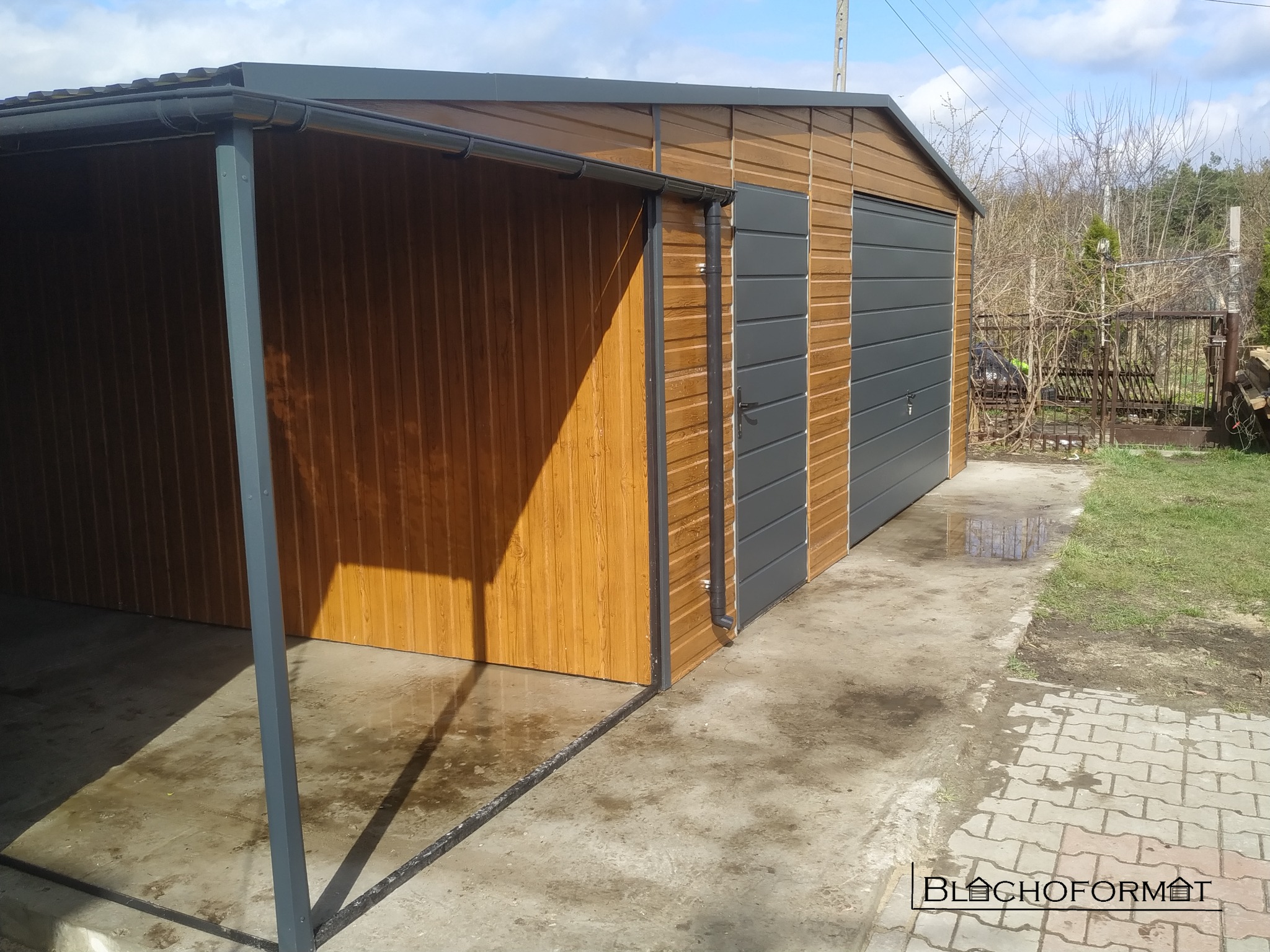 Gotowy garaż blaszany z wiatą, wykończony drewnopodobną blachą, z szarą bramą segmentową i drzwiami, widoczny na betonowym podłożu.