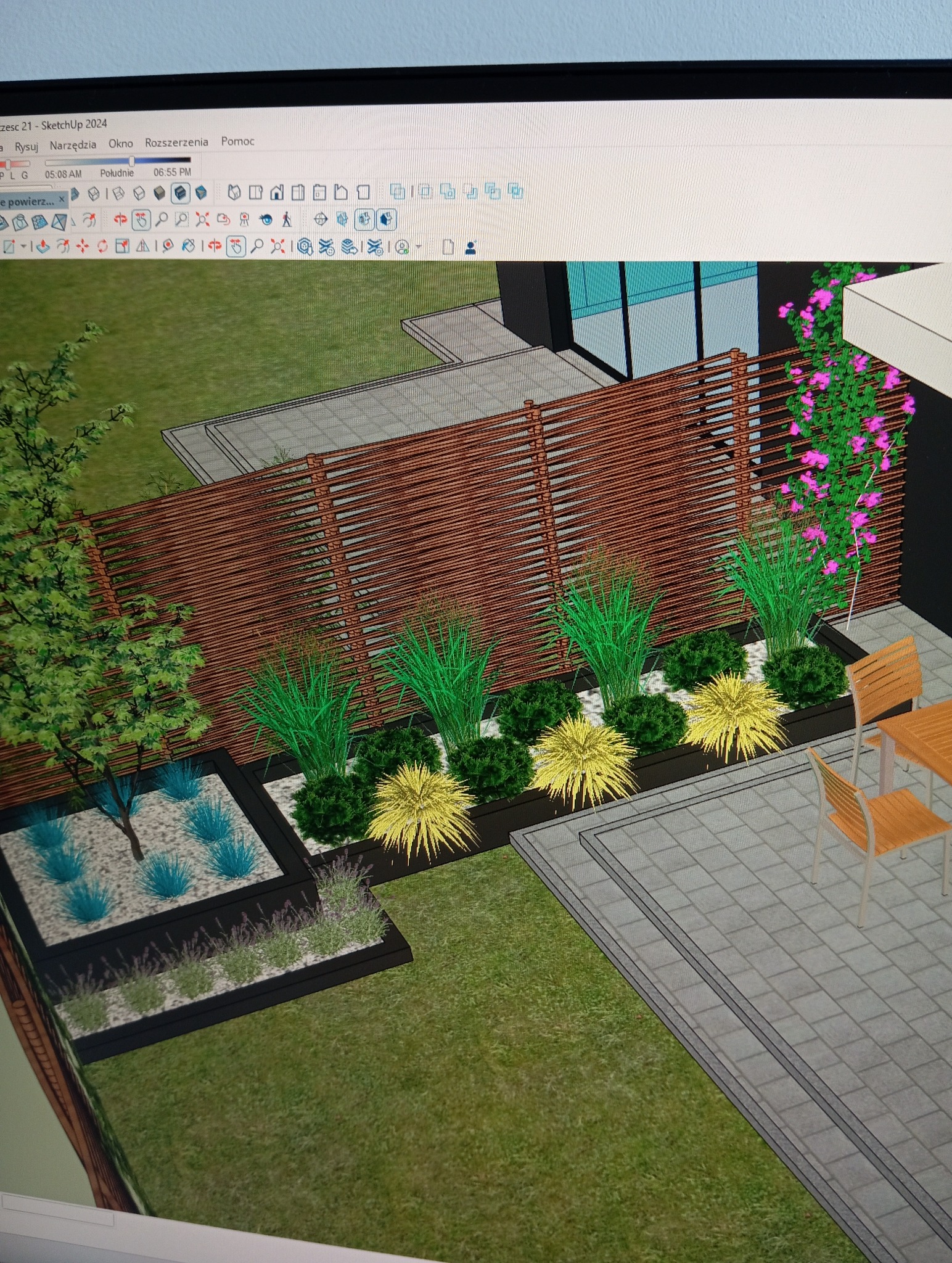 Wizualizacja 3D ogrodu z drewnianym ogrodzeniem, rabatami z ozdobnymi trawami i krzewami, oraz fragmentem tarasu z meblami ogrodowymi. Projekt w programie graficznym.