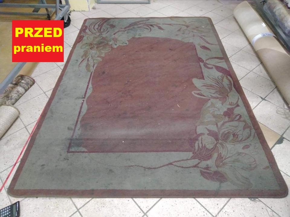Prostokątny, mocno zabrudzony dywan z kwiatowym wzorem przed czyszczeniem, leżący na podłodze wyłożonej płytkami.