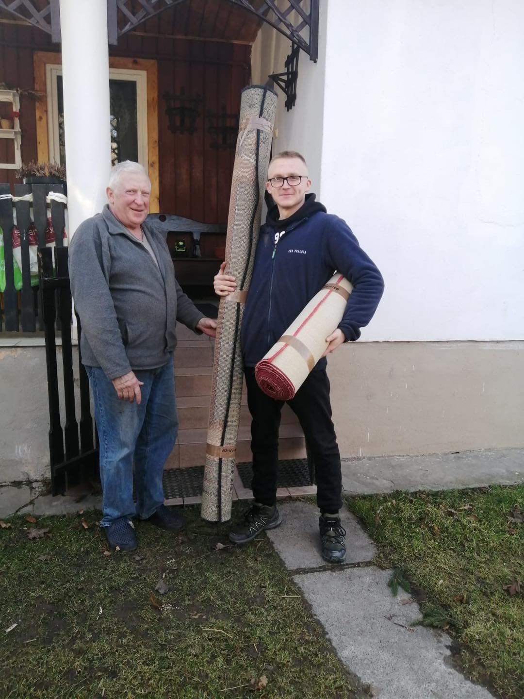 Dwóch mężczyzn stoi przed domem z dwoma zwiniętymi dywanami, jeden z nich trzyma dywany, a drugi opiera rękę na większym dywanie.