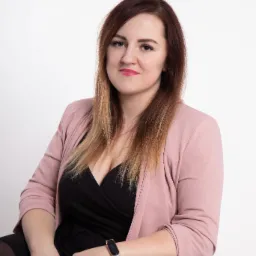 Portret kobiety w różowym żakiecie, siedzącej na czarnym stołku, z zegarkiem na ręce i delikatnym uśmiechem, na jasnym tle.