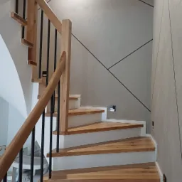 Nowoczesne schody z drewna i metalu z białymi podstopnicami, oświetleniem punktowym i geometrycznym wzorem na ścianie. Ujęcie z dołu, widoczne detale wykończenia.