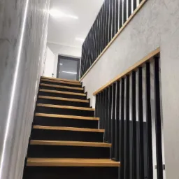 Nowoczesne schody drewniane z czarnymi podstopnicami i metalową balustradą w minimalistycznym wnętrzu z oświetleniem LED na ścianie.