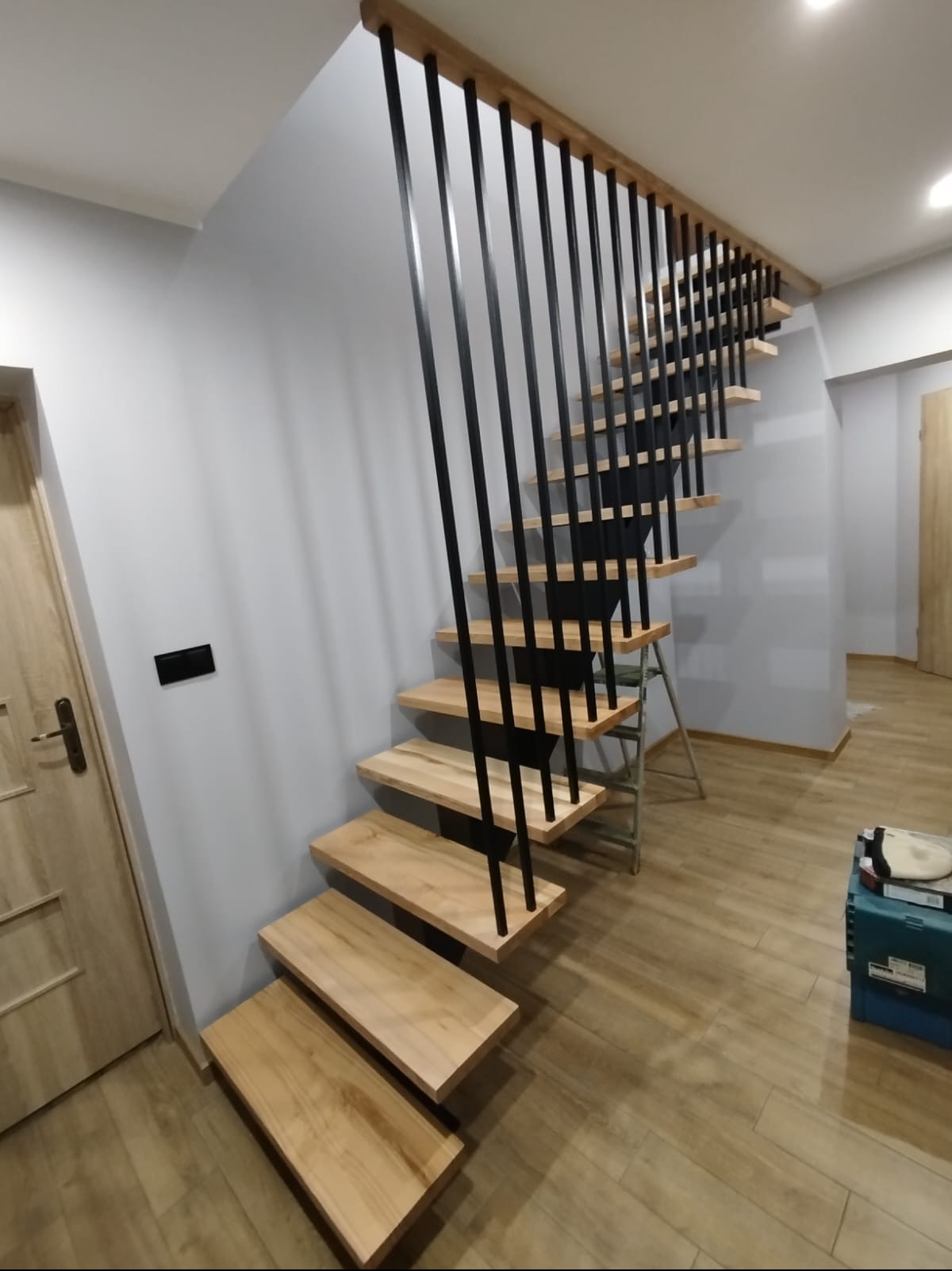 Nowoczesne schody z drewnianymi stopniami i czarnymi, metalowymi balustradami w jasnym wnętrzu. Widoczny fragment drabiny monterskiej i narzędzia.