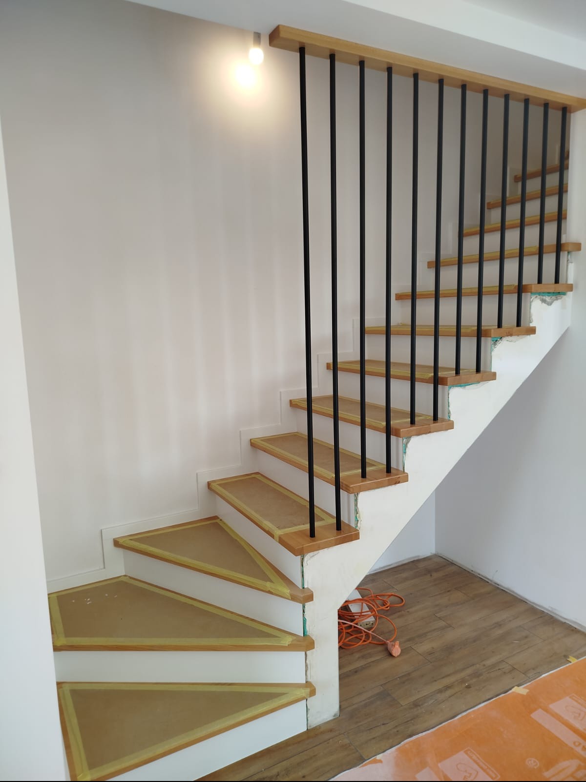 Nowoczesne schody z drewnianymi stopniami i czarnymi, metalowymi tralkami, zabezpieczone taśmą malarską. Widok na jasne wnętrze w trakcie prac wykończeniowych.