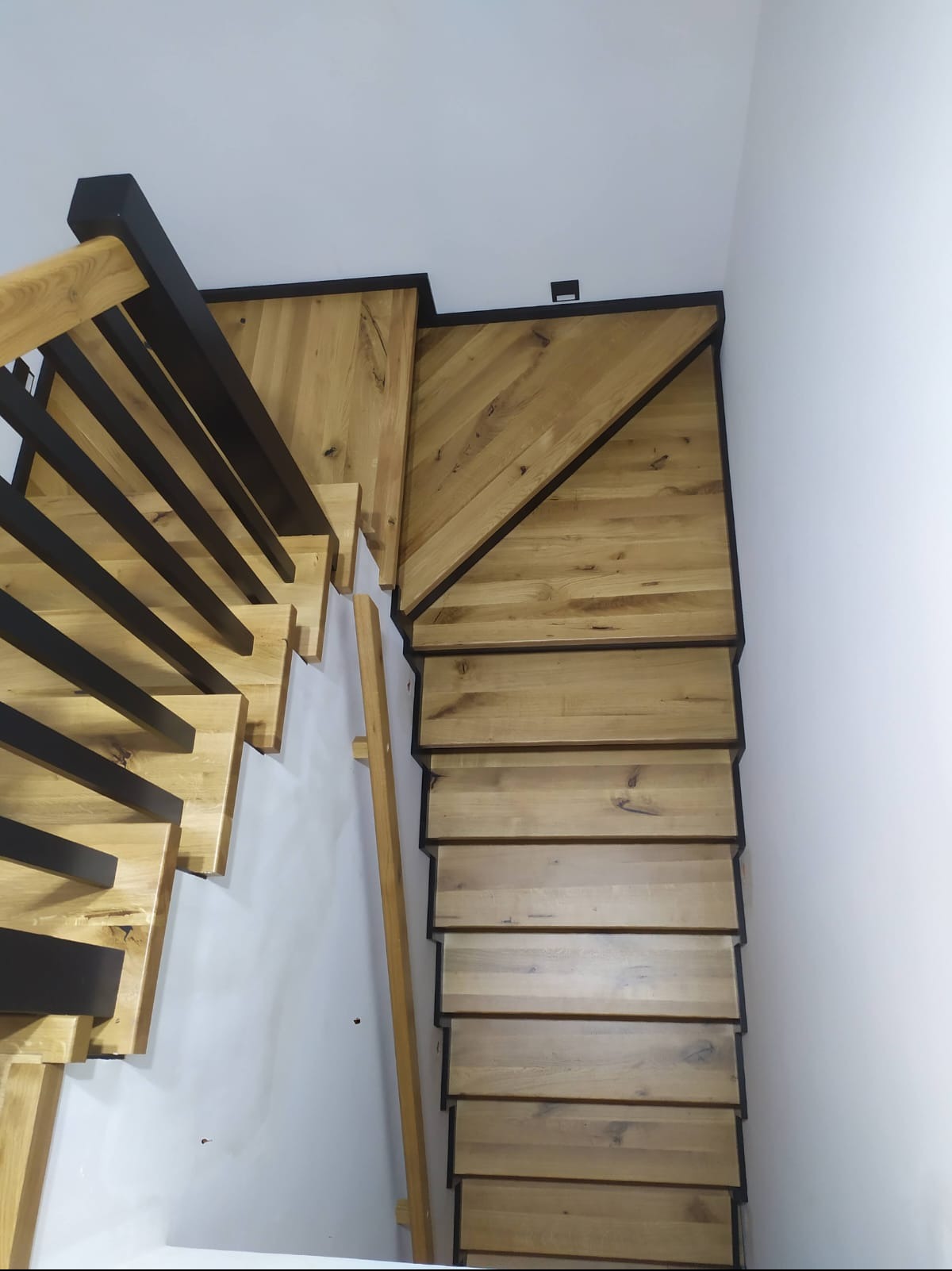 Drewniane schody z czarnymi elementami metalowymi, widok z góry, ukazujący naturalny odcień drewna i geometryczne wzory na podeście przy ścianie.