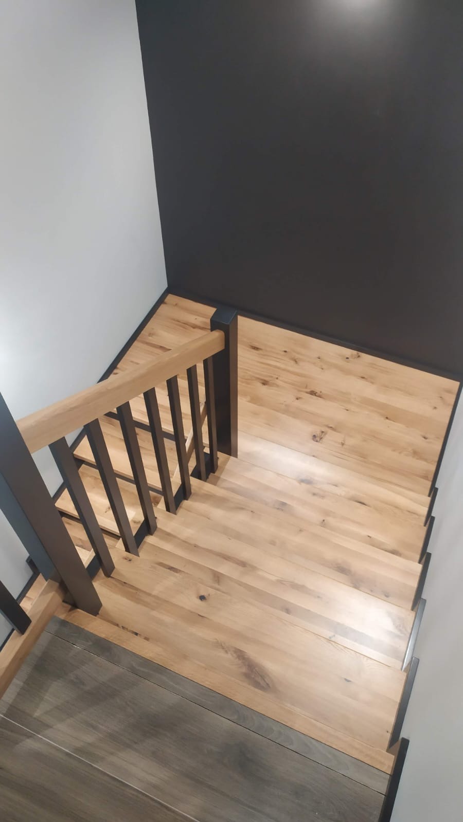 Jasne, drewniane schody z czarną balustradą i podstopnicami, widok z góry, kontrastujące ściany: biała i grafitowa. Minimalistyczny design.