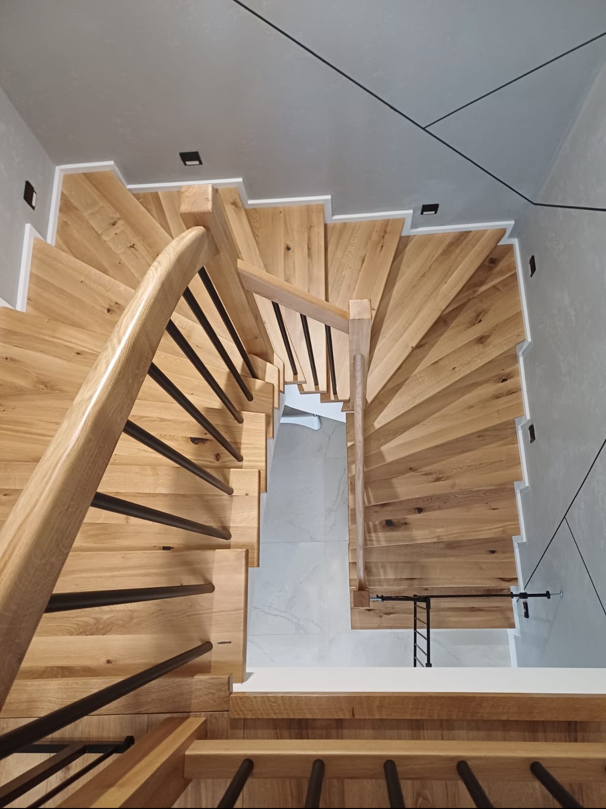 Spiralne schody dębowe z czarnymi balustradami, widok z góry, oświetlenie punktowe. Realizacja w Rawiczu. Unikalny design, nowoczesne wykończenie.