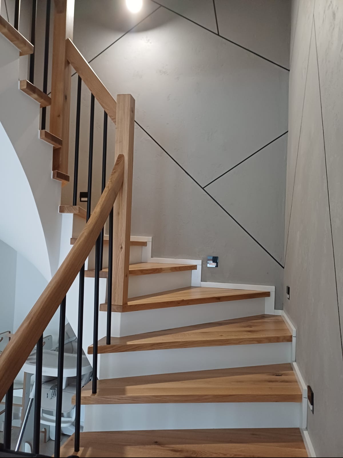Nowoczesne schody z drewna i metalu z białymi podstopnicami, oświetleniem punktowym i geometrycznym wzorem na ścianie. Ujęcie z dołu, widoczne detale wykończenia.