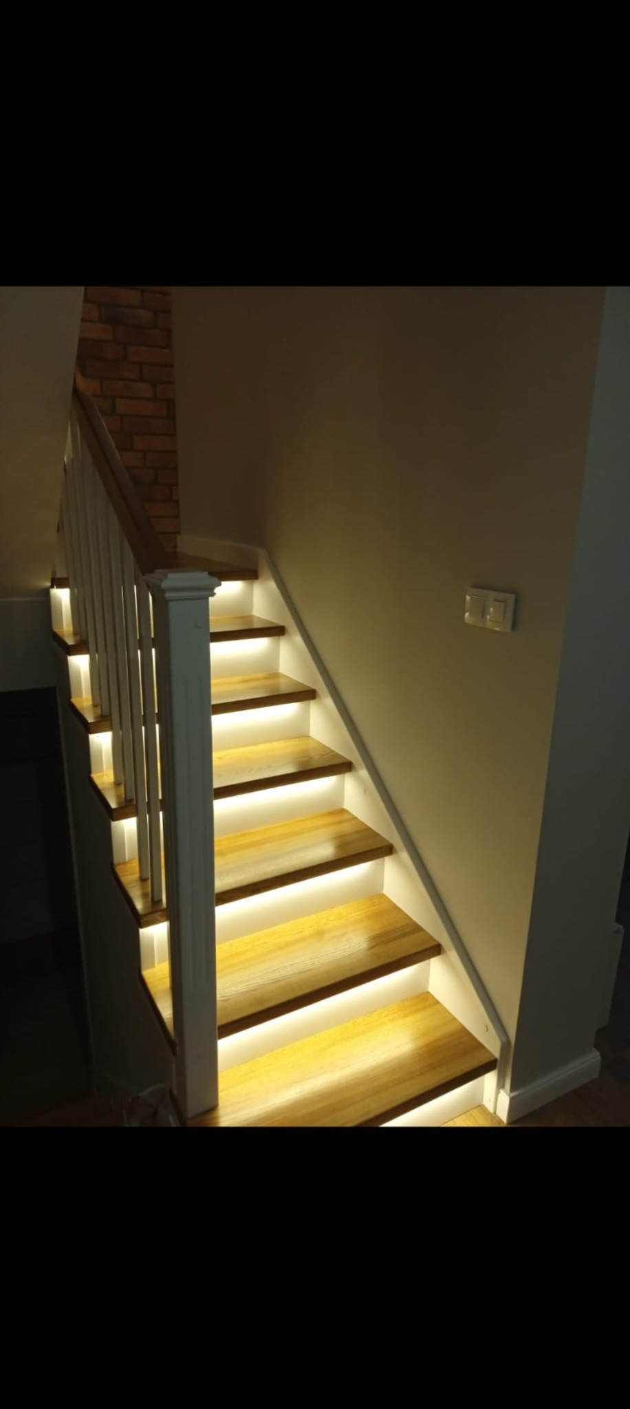 Klatka schodowa z drewnianymi stopniami podświetlonymi od spodu ciepłym światłem LED, biała balustrada, ściana z cegły w tle. Nowoczesny design.