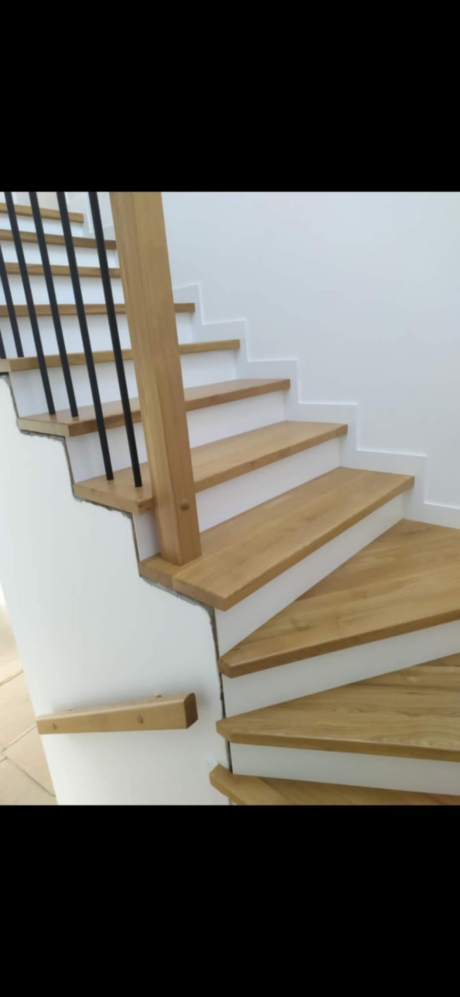 Kaskadowe schody z drewna dębowego z czarną balustradą, osadzone w białej ścianie. Widoczne niedoskonałości wykończenia ściany w miejscu osadzenia schodów.