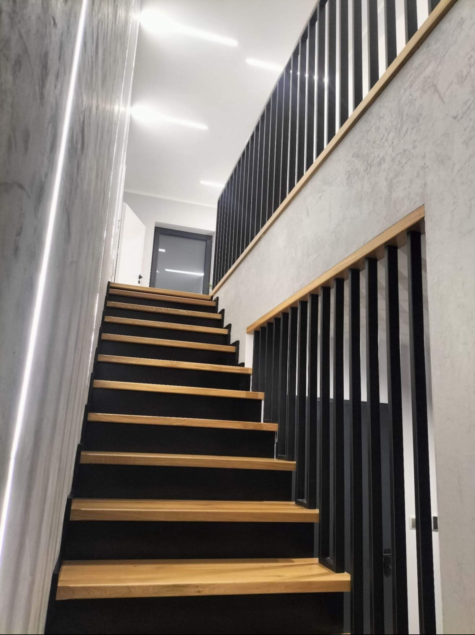 Nowoczesne schody drewniane z czarnymi podstopnicami i metalową balustradą w minimalistycznym wnętrzu z oświetleniem LED na ścianie.