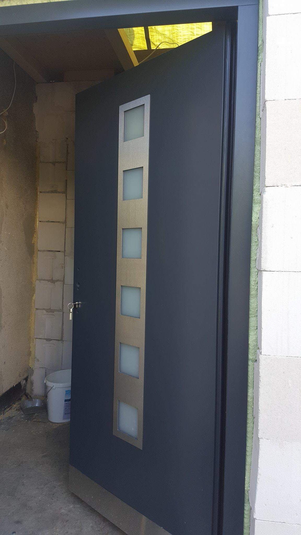 Ciemnoszare drzwi wejściowe z pionowym panelem z pięcioma mlecznymi okienkami, zamontowane w surowym otworze, widoczna piana montażowa i klucz w zamku.