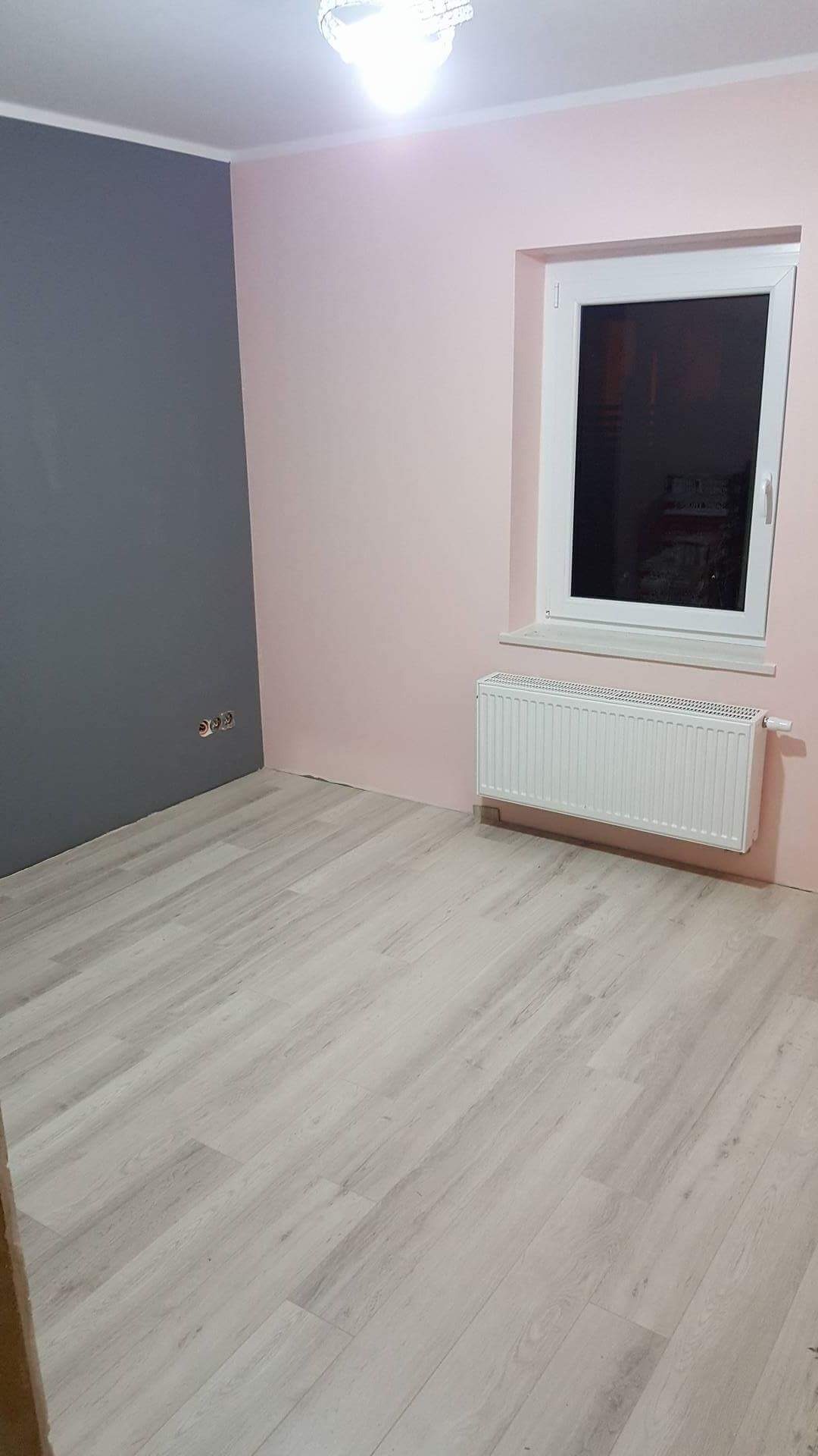 Pokój po remoncie: ściana szara i różowa, panele podłogowe imitujące jasne drewno, biały grzejnik pod oknem, widoczne gniazdka elektryczne na szarej ścianie.
