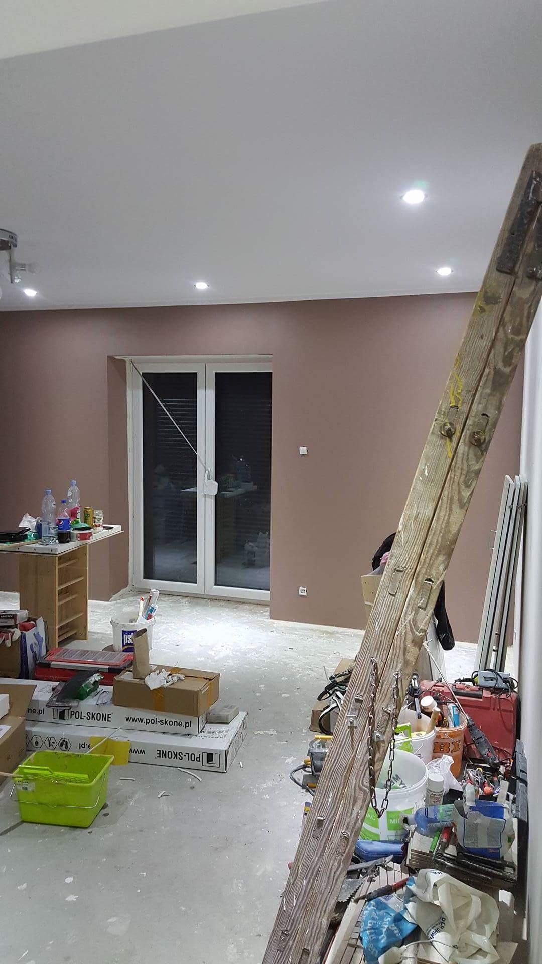 Wnętrze w trakcie prac wykończeniowych: ściany pomalowane na kolor brudnego różu, widoczna drabina, narzędzia malarskie, kartony po drzwiach i okno balkonowe z żaluzjami.
