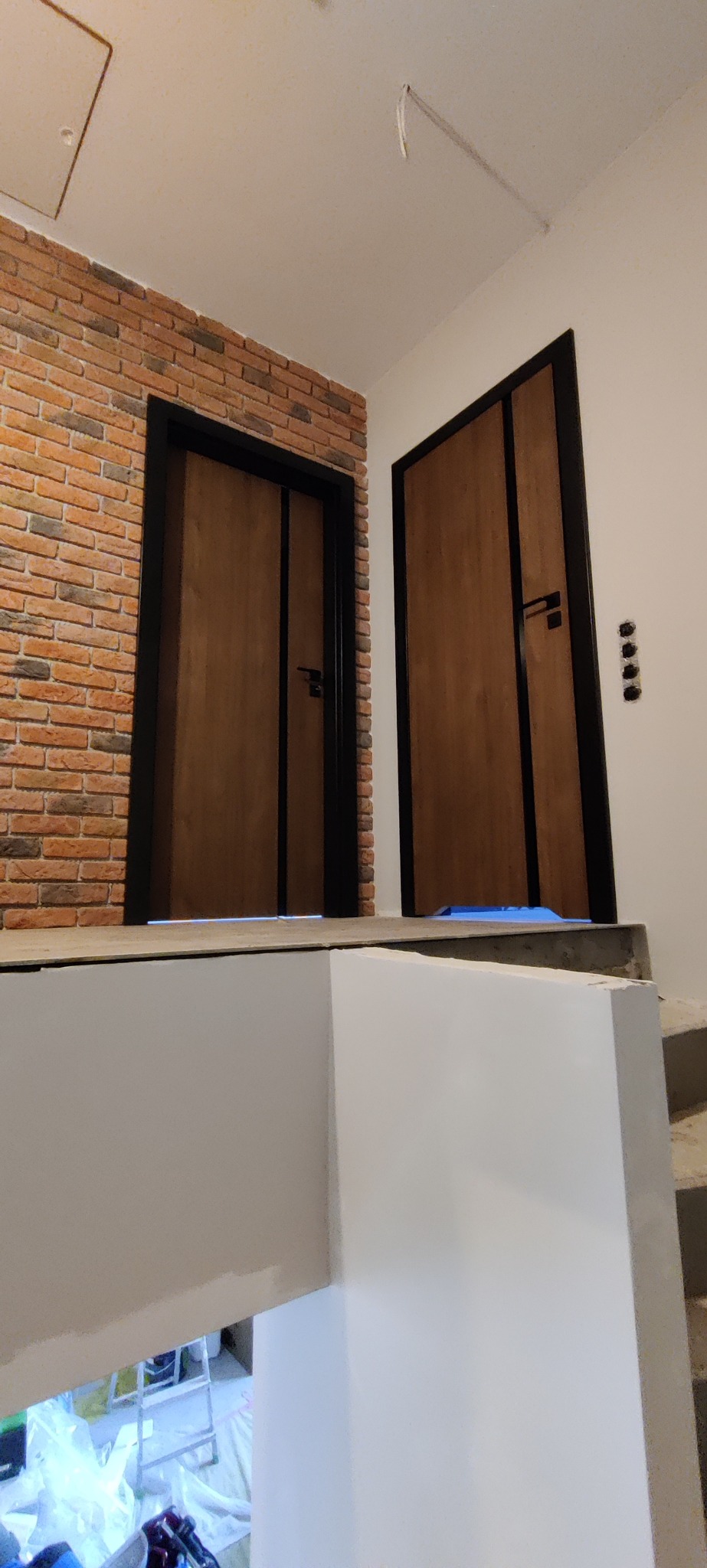 Dwa drewniane drzwi z czarną ramą, jedno przy ceglanej ścianie, drugie przy białej, widoczne schody i zabezpieczenie pod drzwiami.