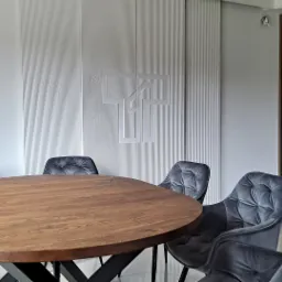 Owalny drewniany stół otoczony sześcioma szarymi, pikowanymi krzesłami na tle białej ściany z logo, widoczne drzwi z drewnianymi lamelami.
