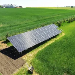 Widok z góry na panel słoneczny umieszczony na skraju pola, obok ogrodzenia z siatki i małego ogródka warzywnego, z taczką w pobliżu.