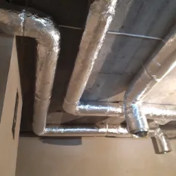 Izolowane rury wentylacyjne, pokryte folią aluminiową, zamontowane pod betonowym sufitem. Widoczne załamania rur i fragment ściany w tle.