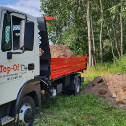 Ciężarówka z czerwoną skrzynią ładunkową wypełniona ziemią, zaparkowana obok pryzmy ziemi na tle drzew, z widocznym logo firmy Top-Of Usługi Minikoparką/Transport.