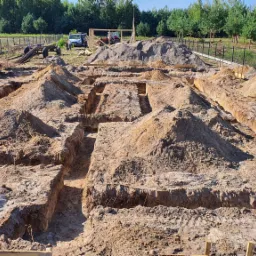 Przygotowanie terenu pod budowę w Jeżowe: wykopane rowy pod fundamenty z widocznymi stertami ziemi, w tle ogrodzenie, drzewa i częściowo widoczny sprzęt budowlany.