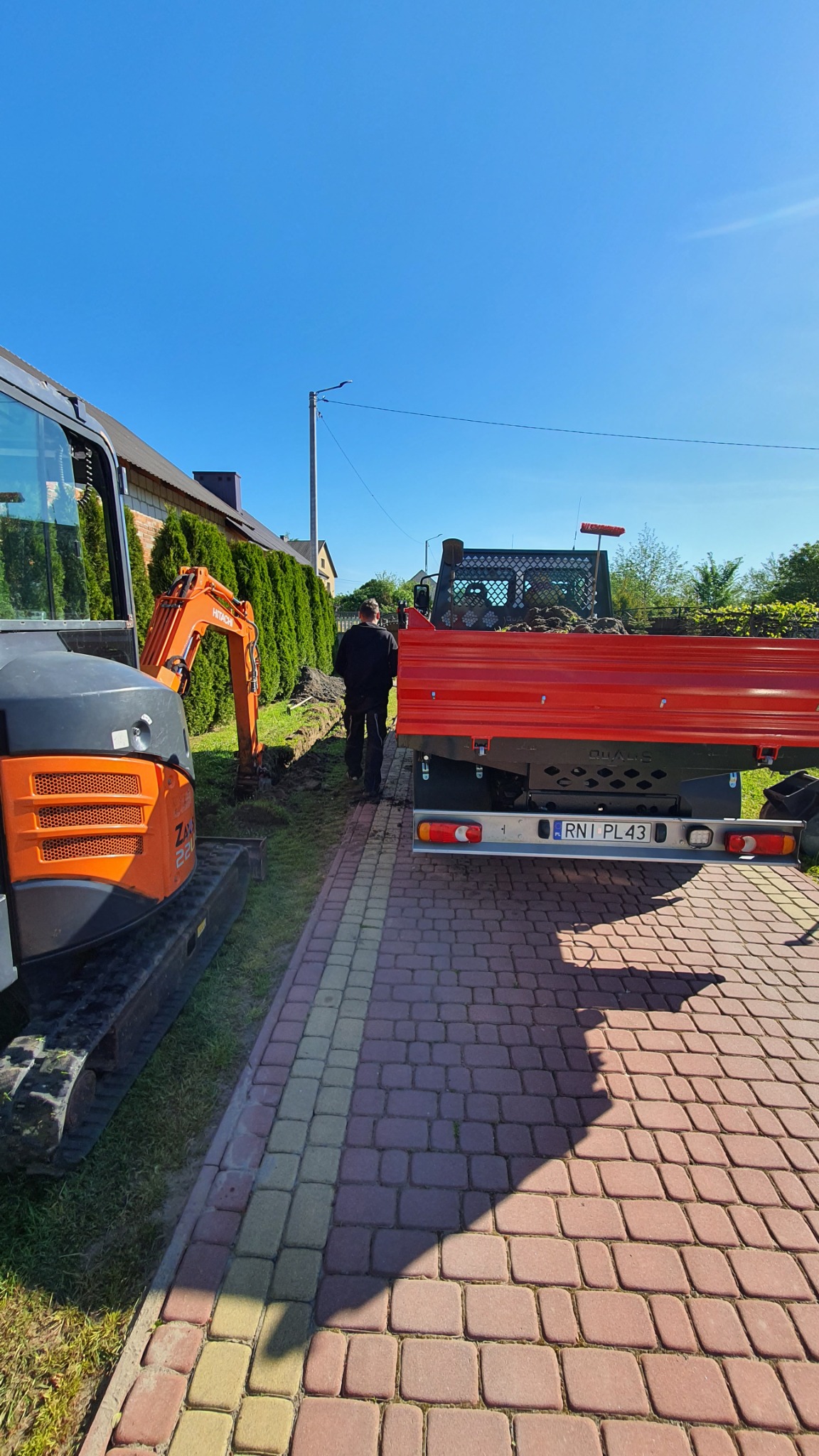Pomarańczowa mini koparka Hitachi Zaxis 26U i wywrotka z ziemią na tle wykopu pod instalacje, widok z perspektywy chodnika z kostki brukowej w słoneczny dzień.