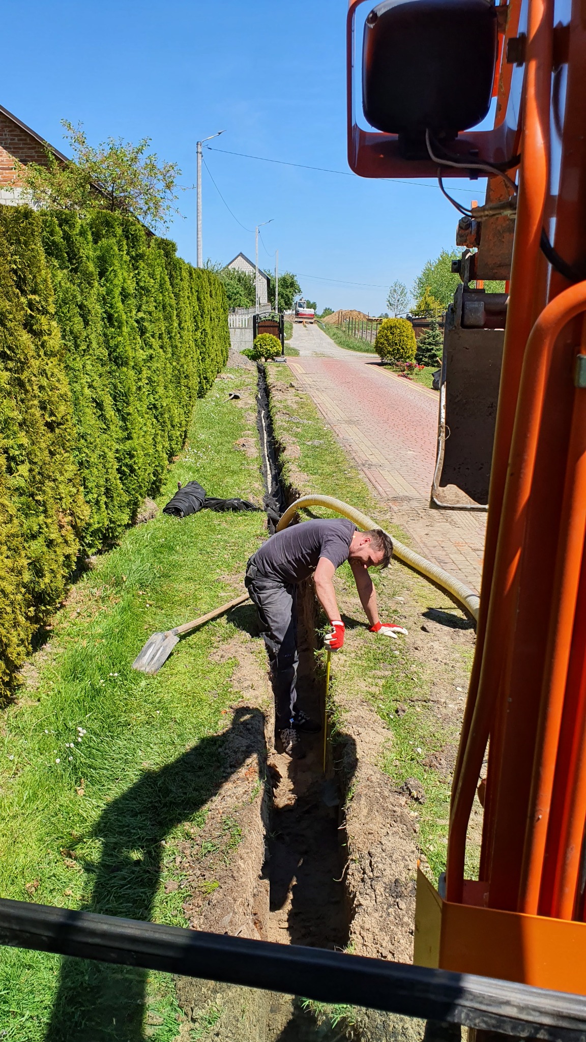 Pracownik w czerwonych rękawicach, pochylony nad wąskim wykopem w trawie, obok leży szpadel, widoczna pomarańczowa maszyna i rura, w tle dom i droga.