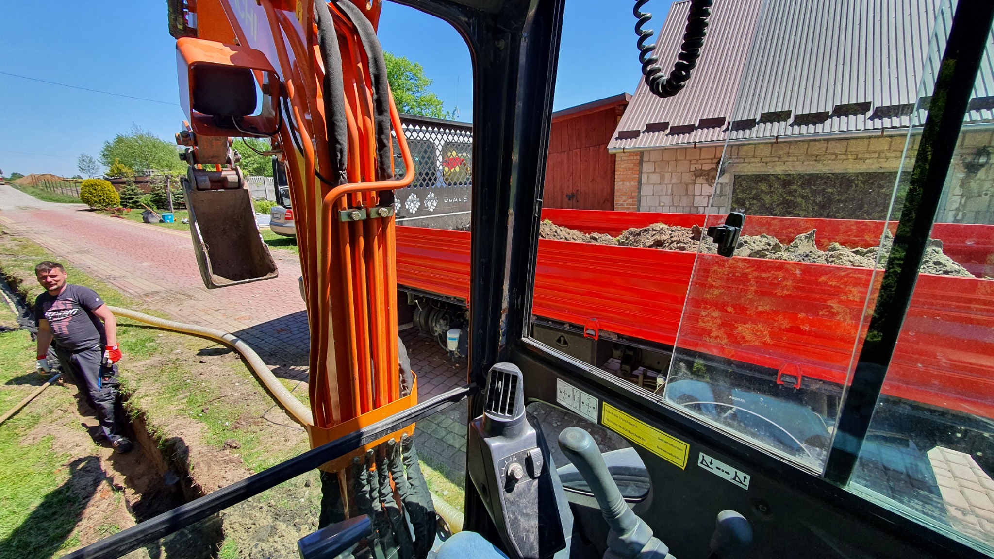 Widok z kabiny pomarańczowej koparki na wykop w trawie, pracownika z rurą oraz na czerwoną ciężarówkę z ziemią w tle.